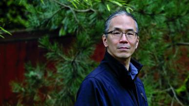 Ted Chiang looks at the camera. He is wearing glasses, a blue coat and is standing in front of green leaf trees. 