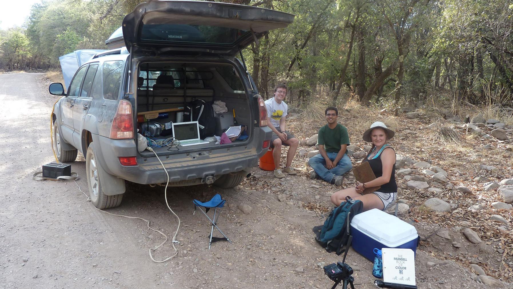 Students sit by an SUV with equipment surrounding them, while conducting fieldwork
