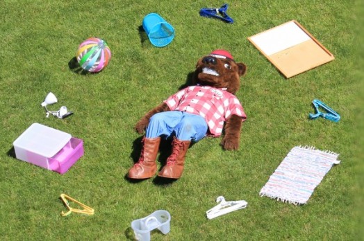 A person in a grizzly bear mascot costume laying on grass surrounded by housewares