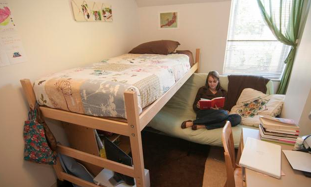 A person reading a book next to a bed in a small dorm room