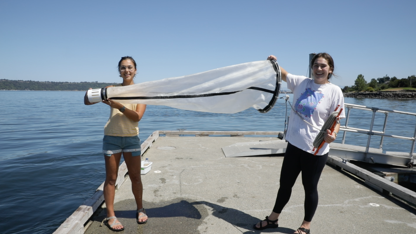 Two female students hold a large water-filtering apparatus on a dock at the Puget Sound