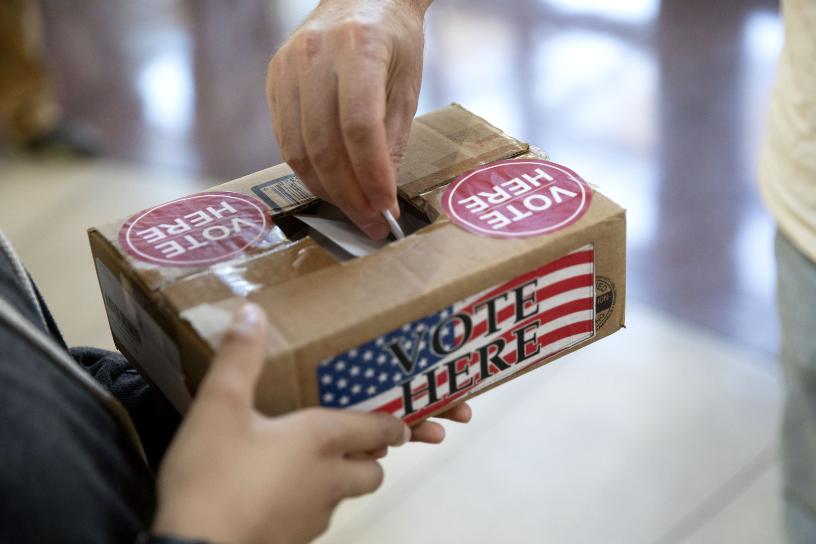 A person holds a box decorated with the stars and stripes, while someone drops a ballot into it from the top.