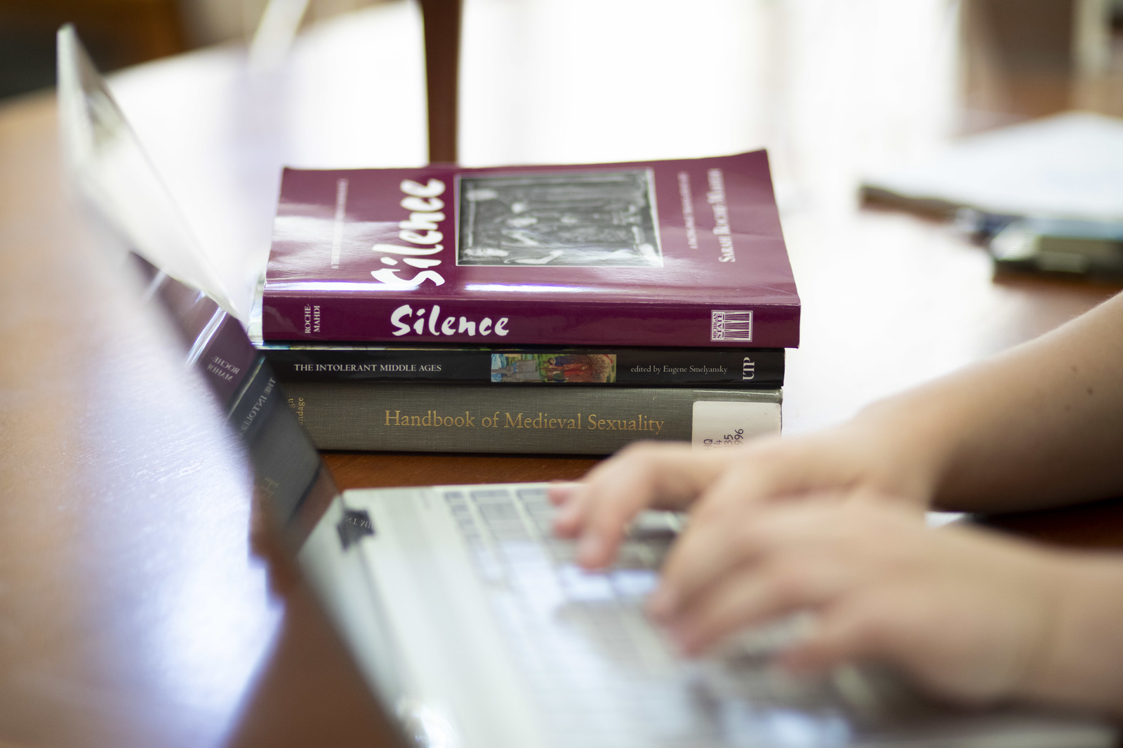 A stack of books sits on a table, visible just beyond two hands typing on a laptop keyboard