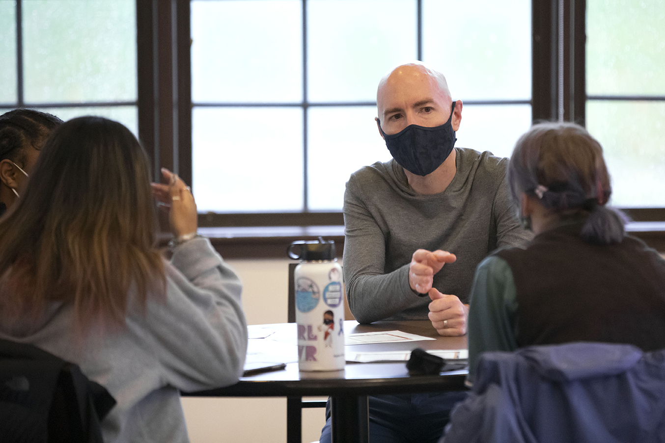 A masked male teacher speaks to students with windows behind him