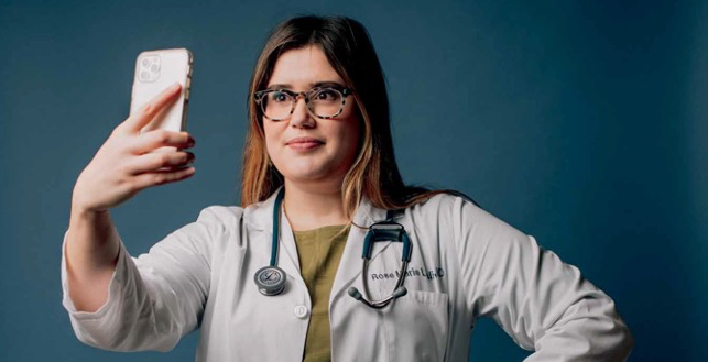 Rose Marie Leslie wearing a lab coat with a stethoscope around her neck, and looking intently at her cell phone. 