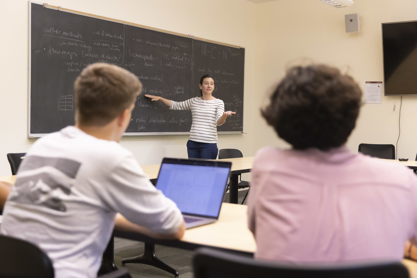 Prof. Courtney Thatcher stands at the chalkboard during a class