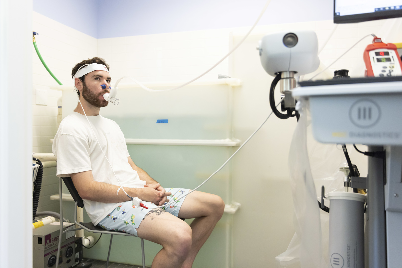A student in a white shirt and swim trunks sits next to a translucent tub of water and breathes into a tube to measure his oxygen levels.
