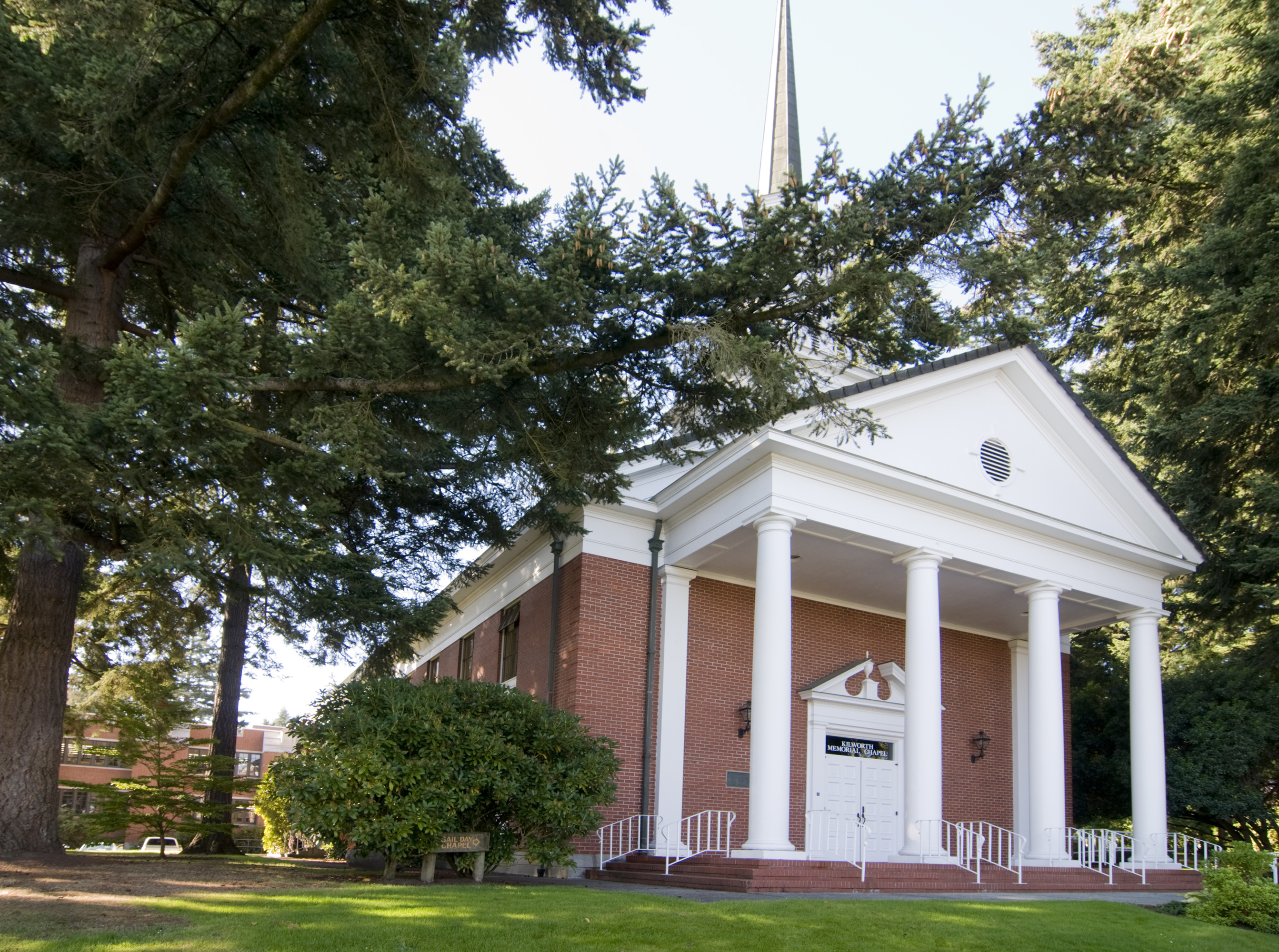 A chapel sits between evergreen trees, with white columns and a steeple. 