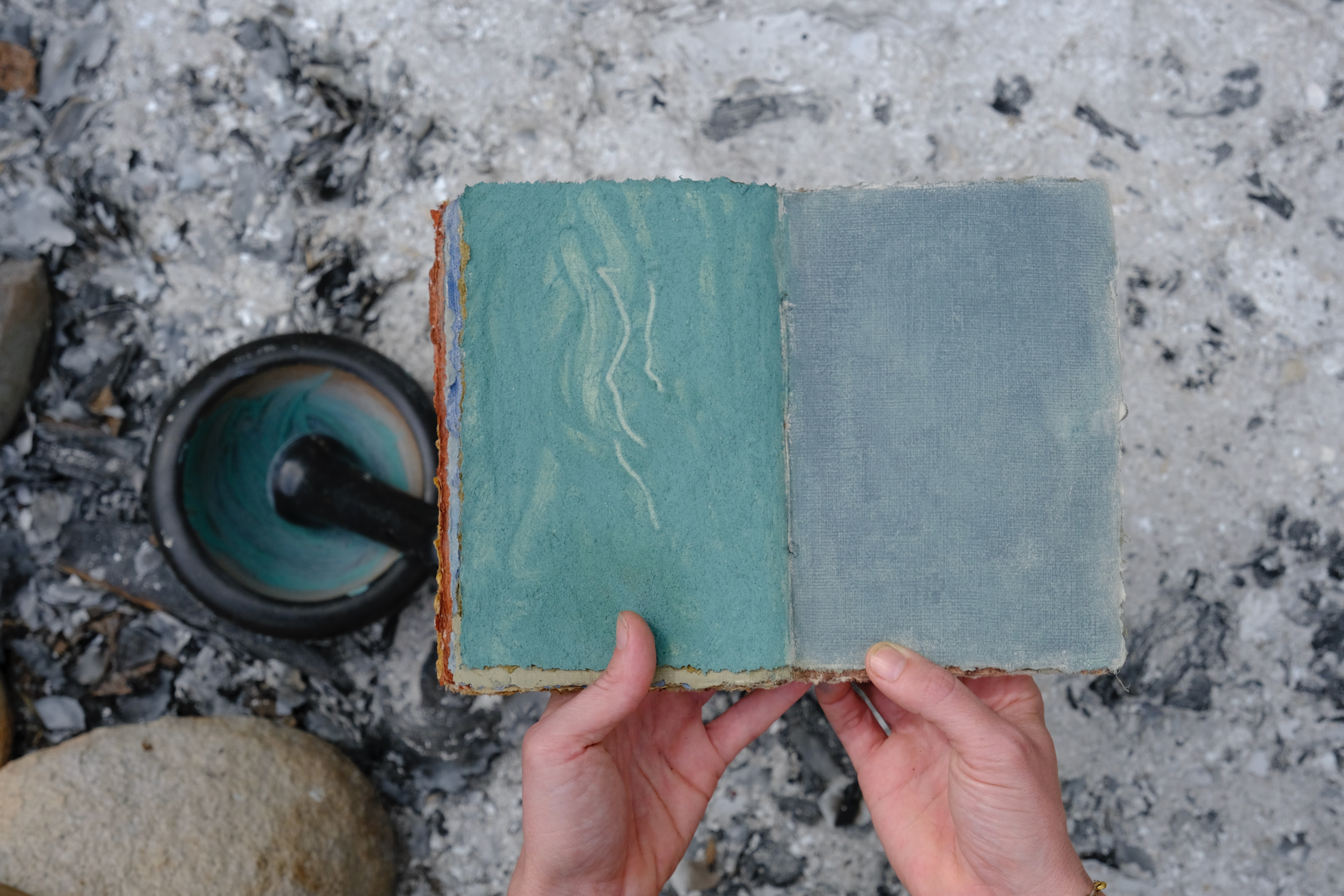 A book with blue pages is opened next to a mortar and pestle of blue powder. 
