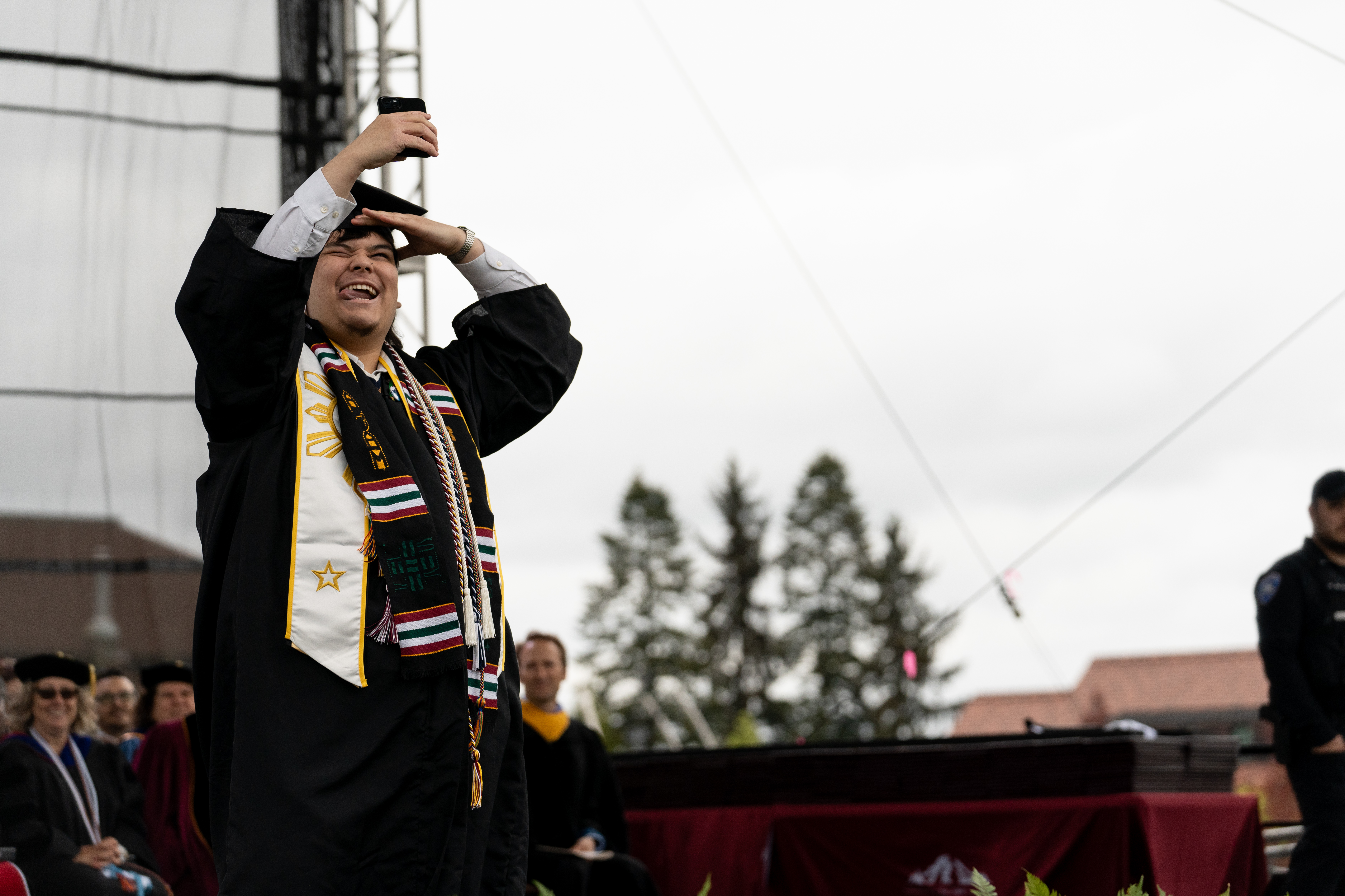 Riley Ofrecio takes a photo of himself with a phone while on the commencement stage.
