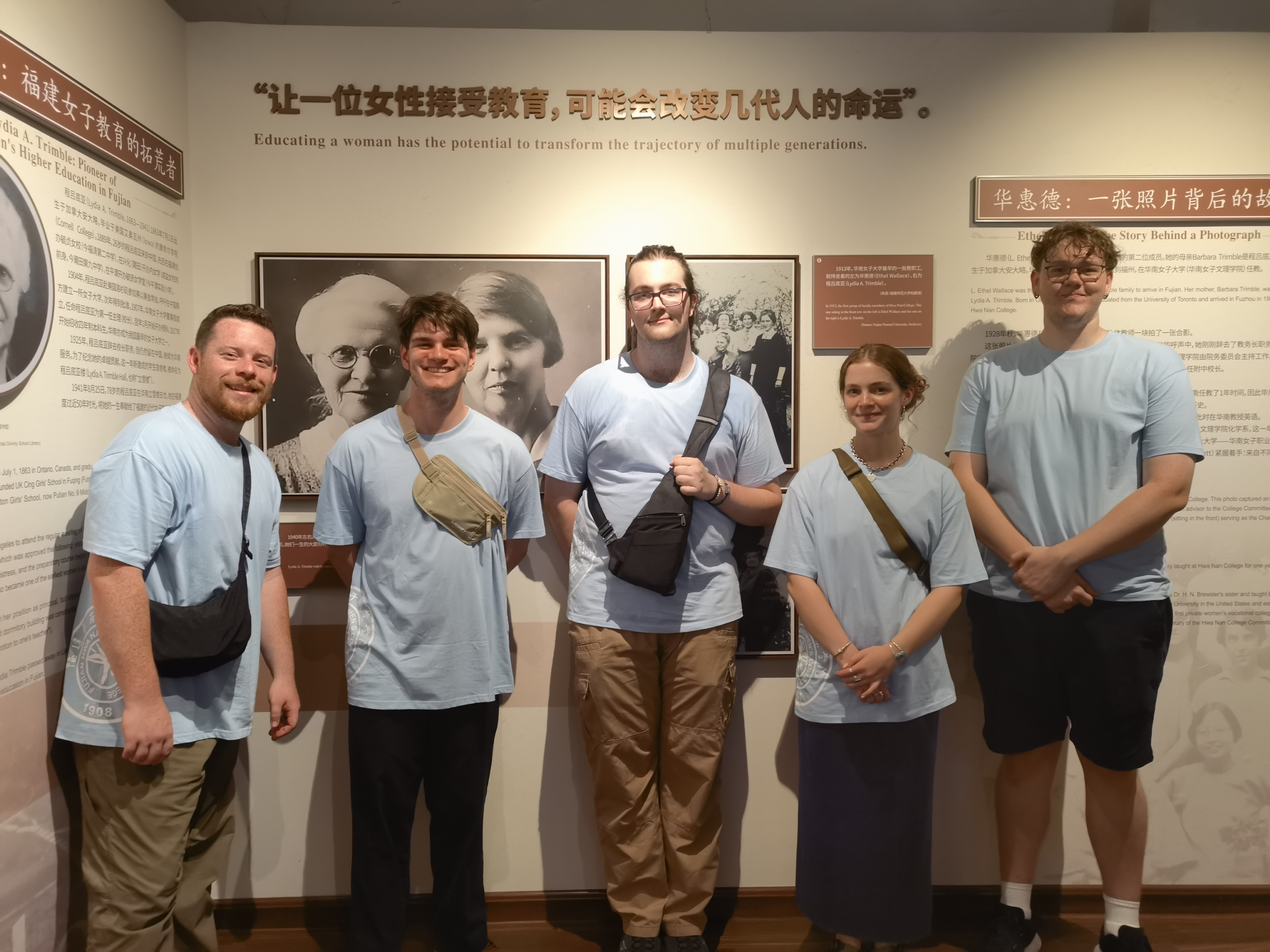 Five students in blue shirts stand in front of a wall with the Trimble family history on it.