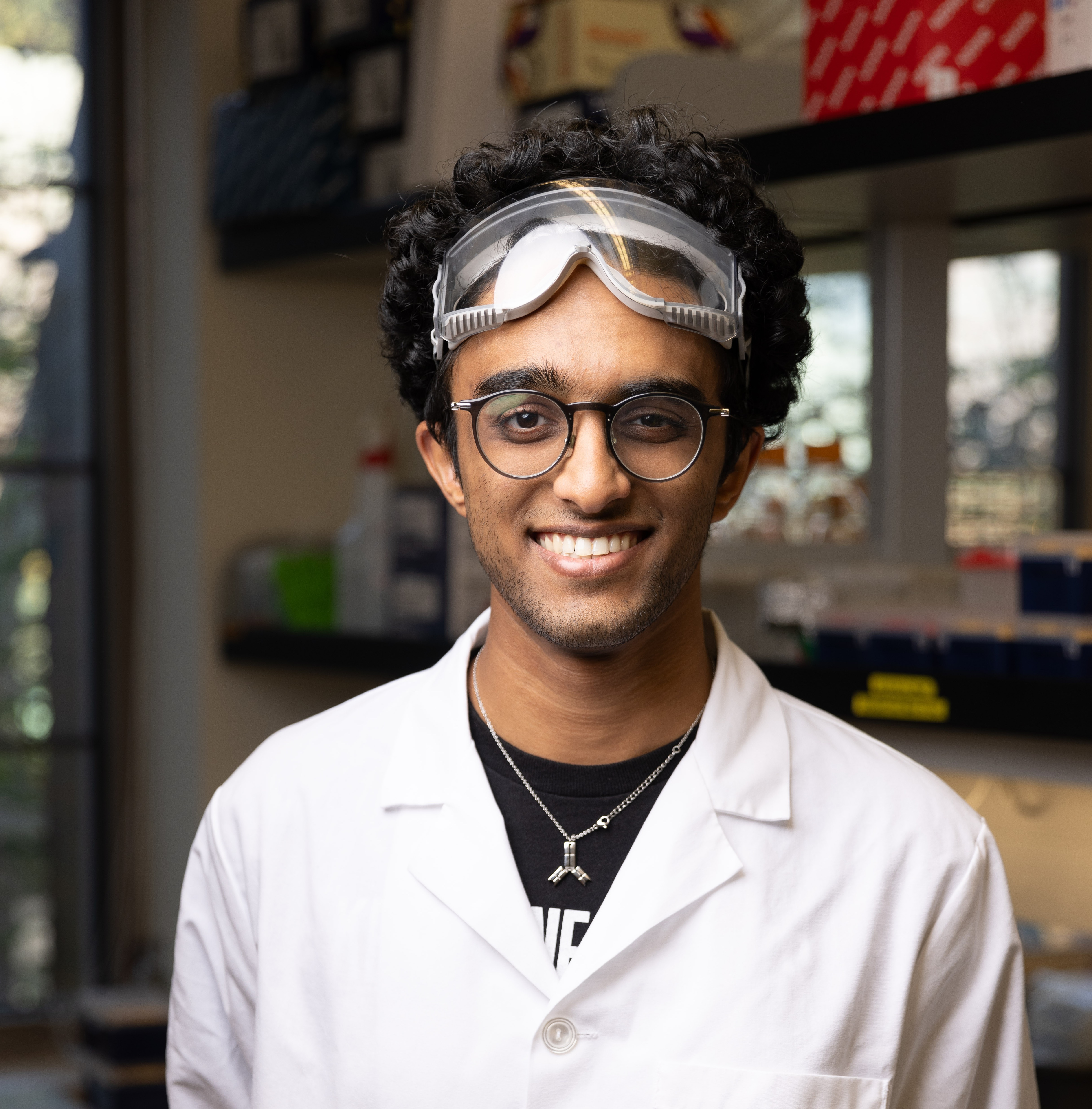 Ishaan wears a white lab coat and goggles in the Chem lab.