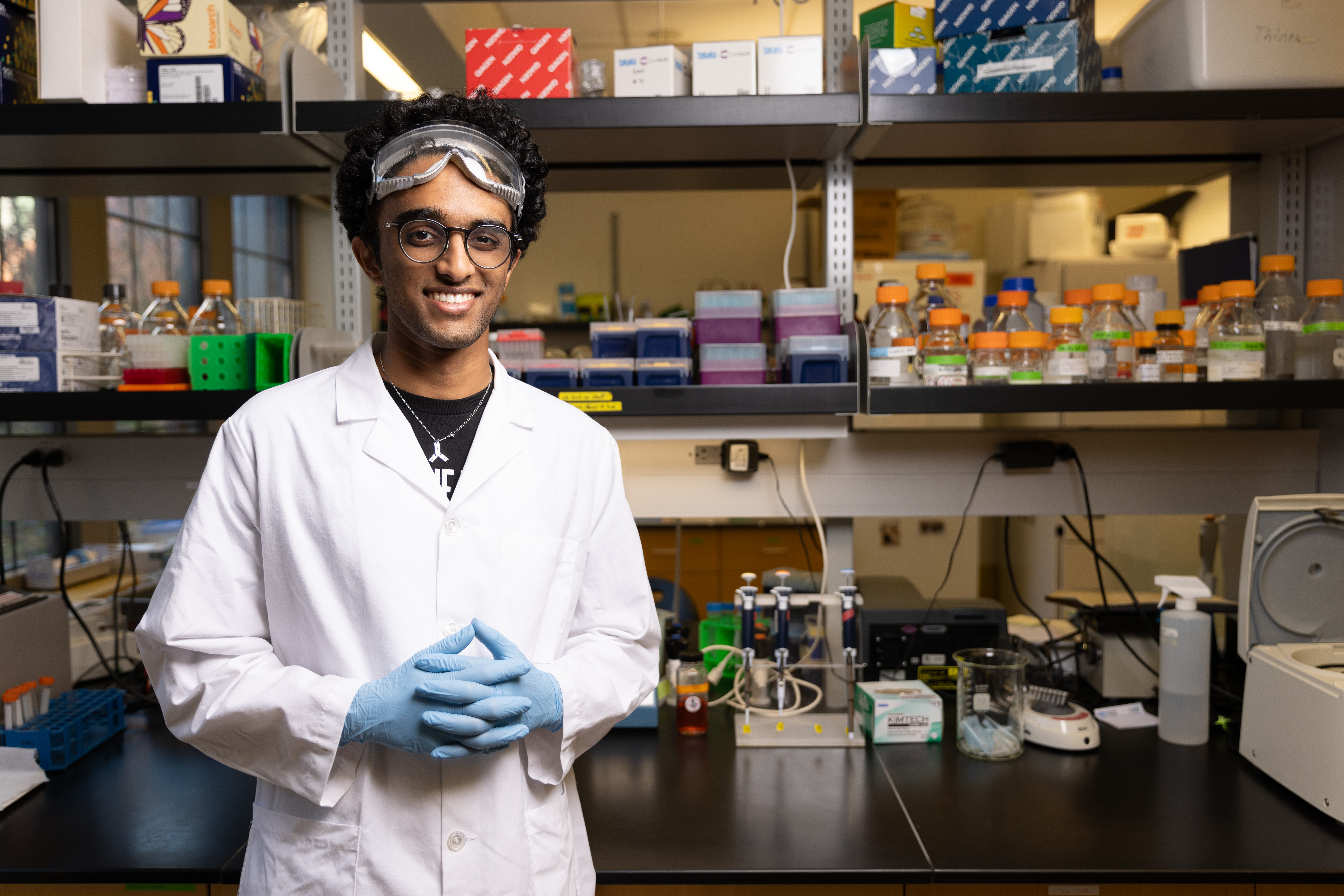 Ishaan Gollamudi stands in a white lab coat, blue rubber gloves and goggles in the chem lab.