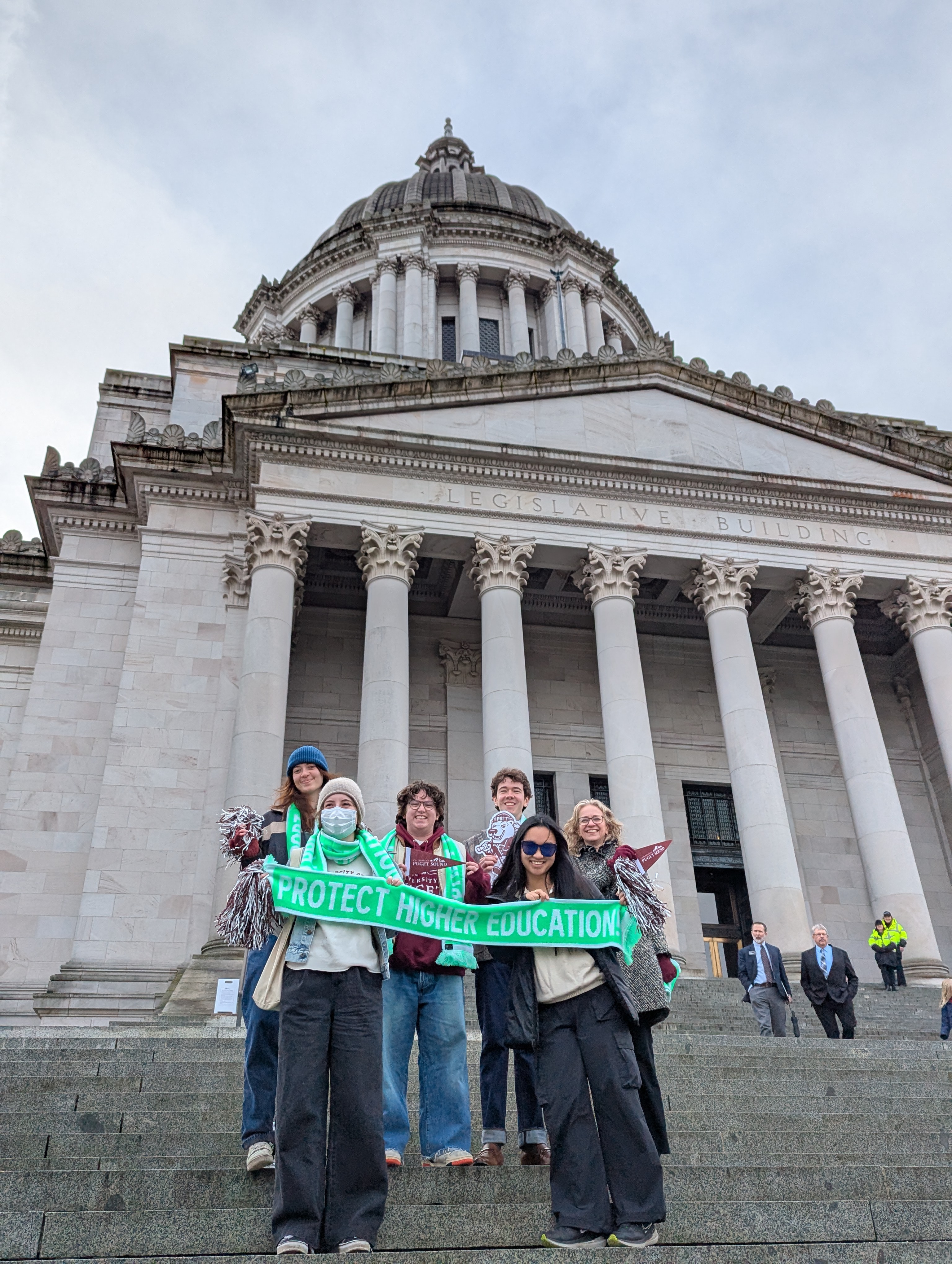 Five students and Prof. Robin Jacobson stand on the Capitol steps holding a green scarf with the words "Protect Higher Education" on it. 