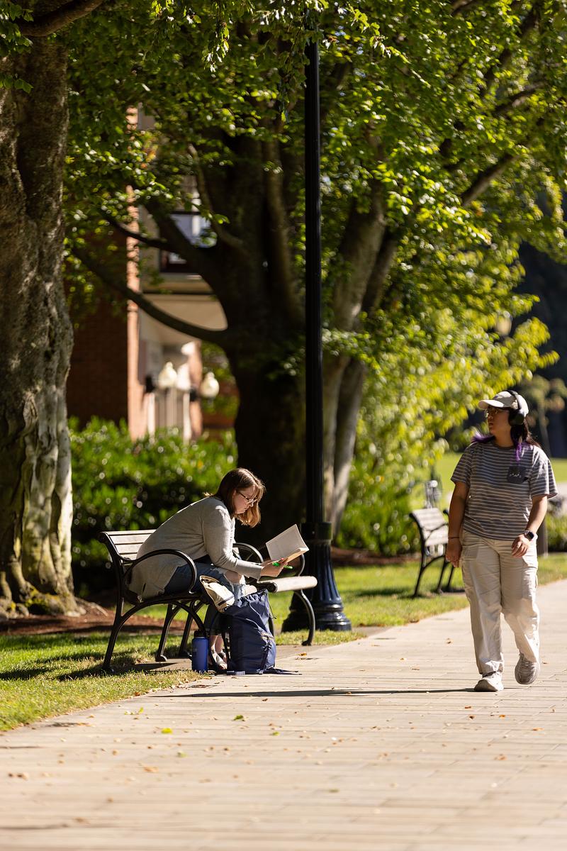 A woman sits on a bench reading a book while another person walks past her listening to headphones. 