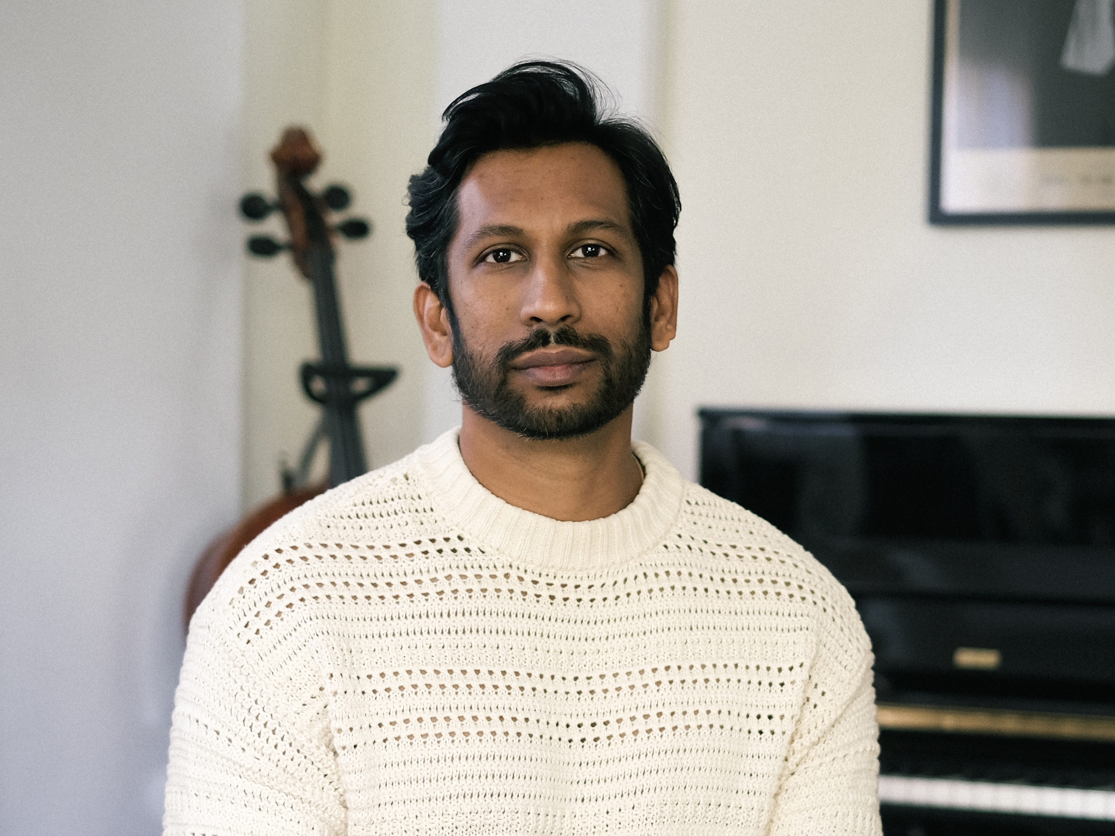 A man with dark hair and a beard sits with a piano and cello behind him. 