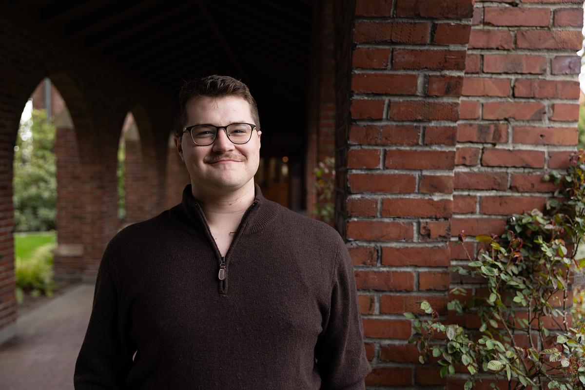 Andrew Benoit stands in front of the red brick arches in a brown sweater.