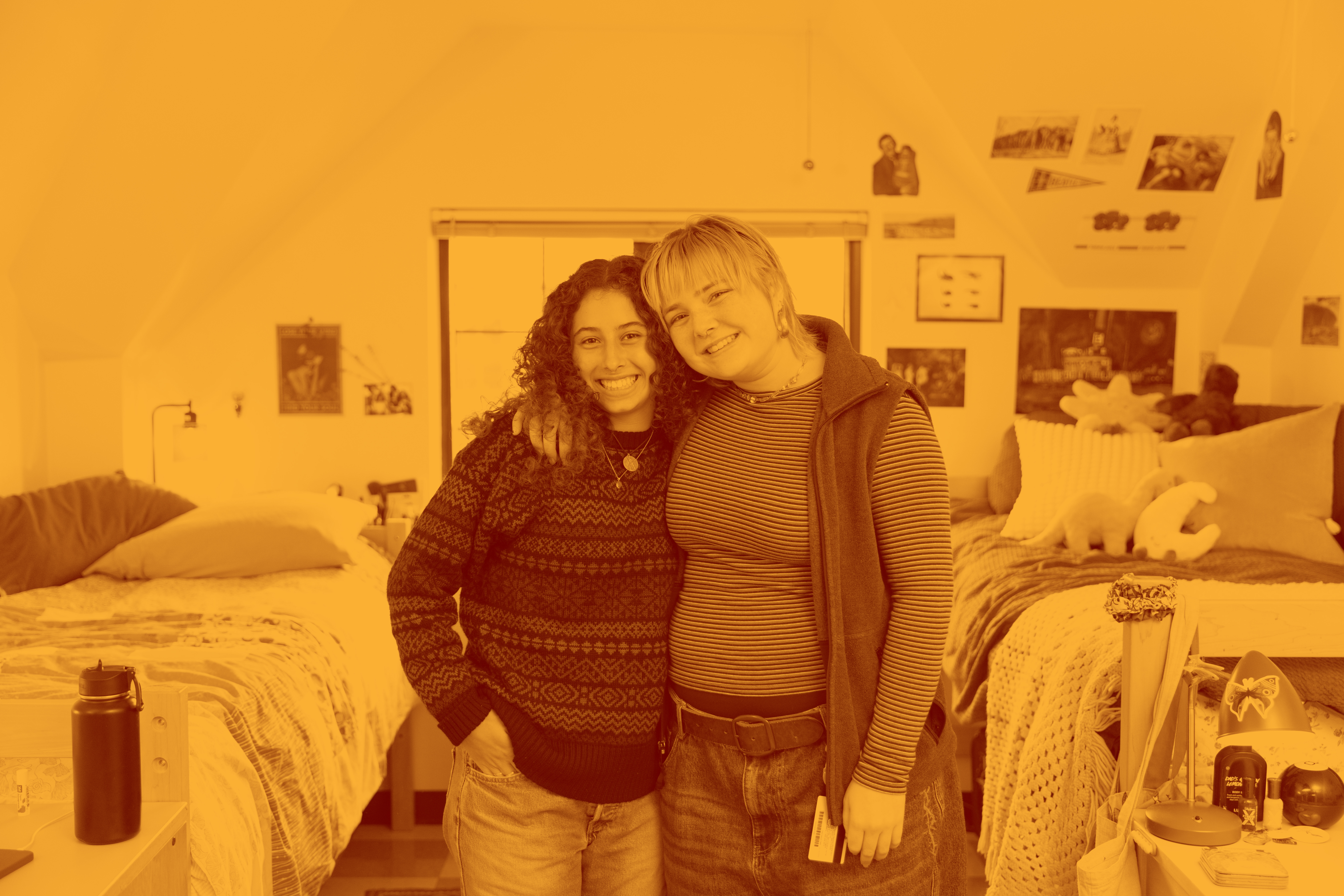 Maroon and yellow tonal image of two students side-hugging in their room.