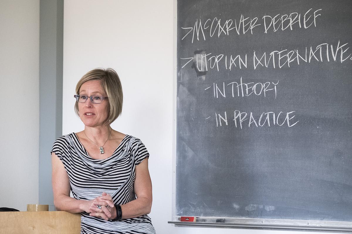 Monica DeHart stands at a podium in front of a chalkboard