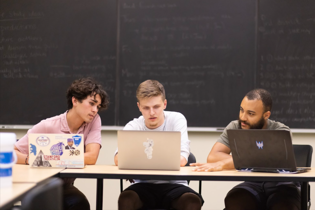 Three students collaborate on laptops in a classroom.
