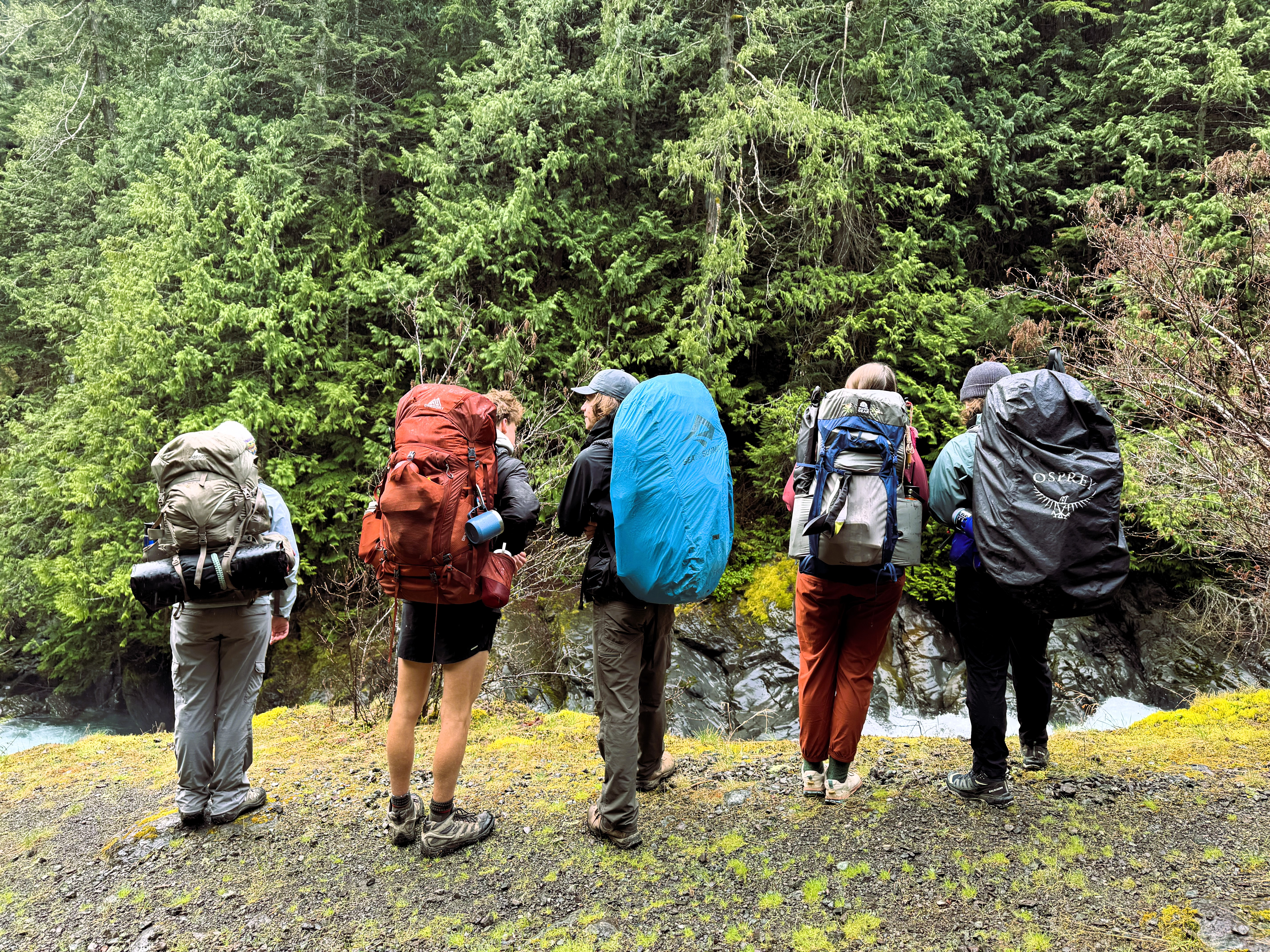 5 backpackers stand on a ledge facing away from the camera looking down on a river that is partially obscured from view.