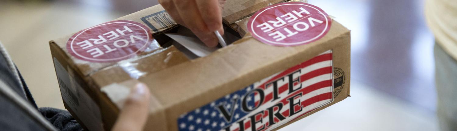 A person holds a box decorated with the stars and stripes, while someone drops a ballot into it from the top.