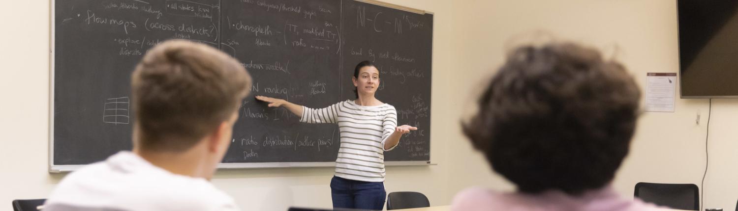 Prof. Courtney Thatcher stands at the chalkboard during a class