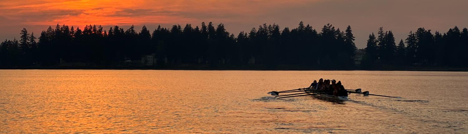 A crew boat is silhouetted by a sunset on a lake with trees in the background