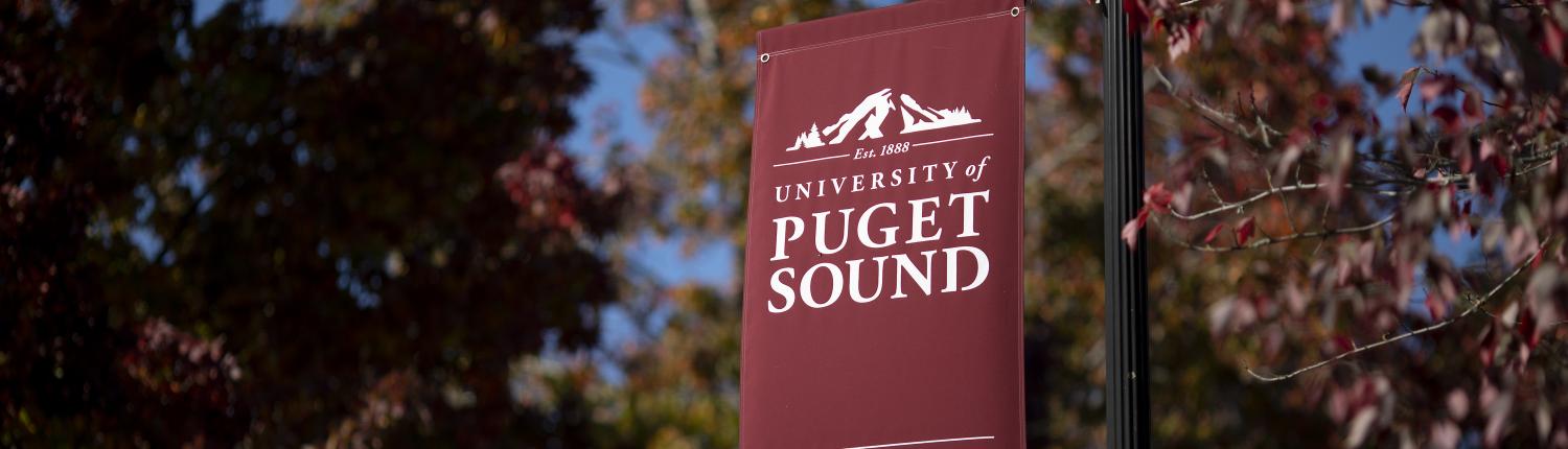 A maroon banner hangs vertically in front of red leaves with the words "University of Puget Sound, To the heights!" displayed. 