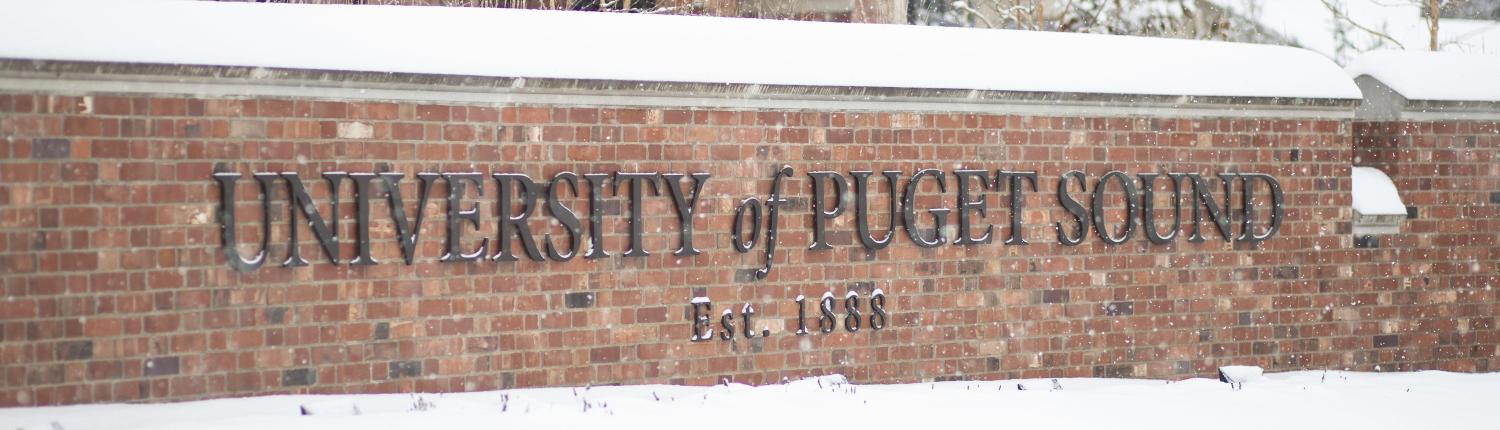 A brick University of Puget Sound sign sits covered in snow. 