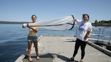 Two female students hold a large water-filtering apparatus on a dock at the Puget Sound