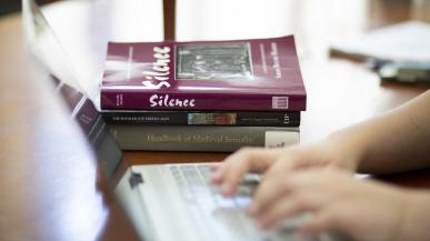 A stack of books sits on a table, visible just beyond two hands typing on a laptop keyboard