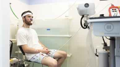 A student in a white shirt and swim trunks sits next to a translucent tub of water and breathes into a tube to measure his oxygen levels.