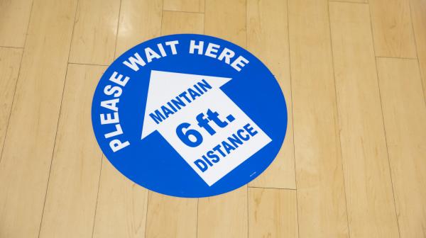A blue and white circular floor sticker that reads "Please wait here" and encourages people to maintain a 6-foot social distance