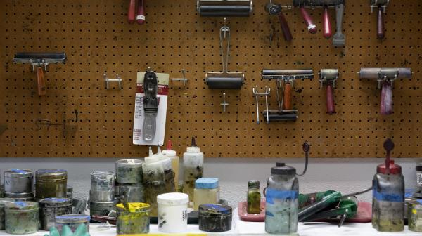 Bottles of colored ink are scattered on a work station with various tools hanging from a pegboard on the wall behind the table