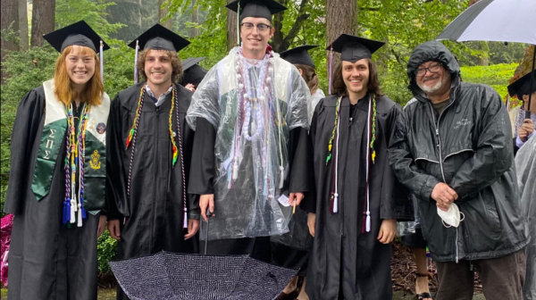 Four students are standing in graduation gowns and rain gear with one other person in a raincoat. There are many trees behind them.