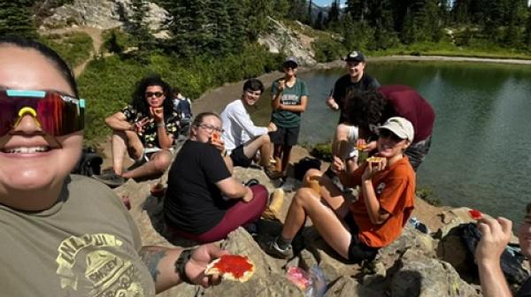 A student in sunglasses takes a selfie with a group of students seated on a rocky area near a lake, eating lunch in the background.