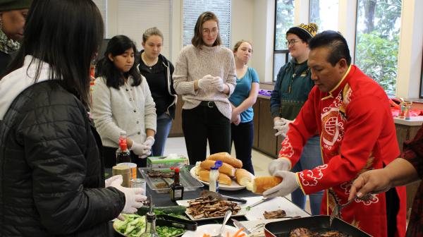 Students gather around a serving table and a man dressed in red traditional Vietnamese-style clothing prepares food. 