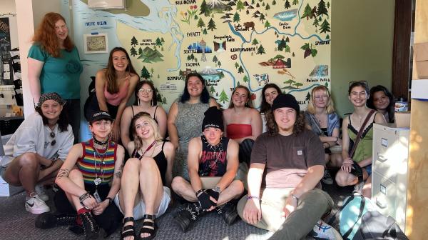 A group of students squish in to be photographed in a smallish room in front of a mural of Washington state.