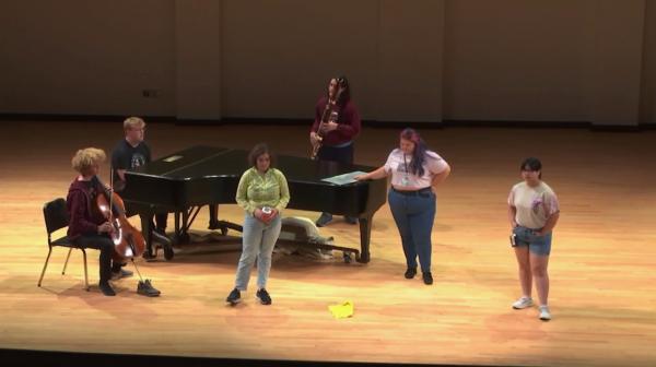 Students on a stage with a piano and additional musical instruments.