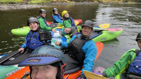 Students in helmets and kayaks float together on a river and smile at the camera.