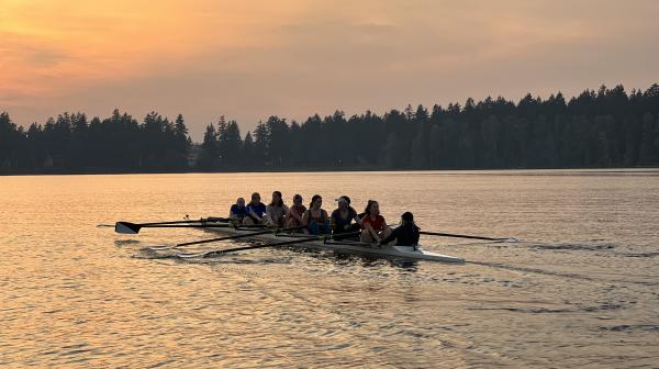 Students row in a crew row boat on a lake during a sunset.