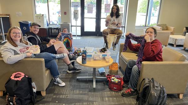 Students sit on couches and chairs with knitting, crochet, or embroidery projects.