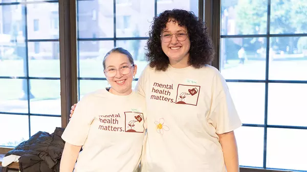 Two students side hugging and smiling at the camera with mental health matters shirts on.