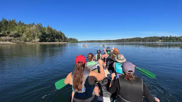 from the perspective of the canoe steerer, 6 people paddle a large canoe with two kayak in from of them in the distance. Trees line the background.