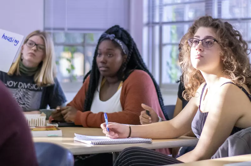 Image of three students in AFAM 101. Student on the far left is holding up a white board sign that says public housing, highlighting a need in a community during an in-class discussion. 