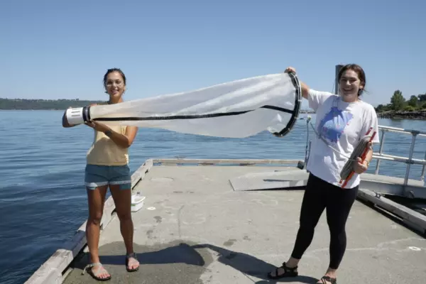 Two female students hold a large water-filtering apparatus on a dock at the Puget Sound
