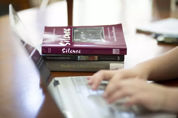 A stack of books sits on a table, visible just beyond two hands typing on a laptop keyboard