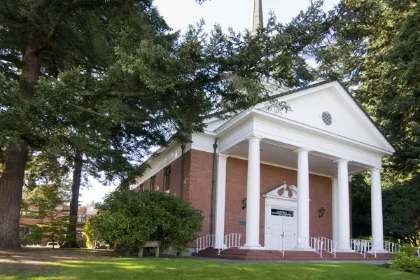 A chapel sits between evergreen trees, with white columns and a steeple. 