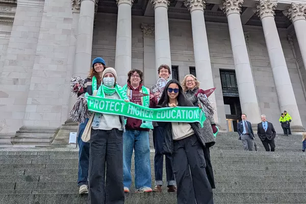 Five University of Puget Sound students and Prof. Robin Jacobson stand on the steps of the Washington State Capitol holding a green scarf with the words "Protect Higher Education" on it. 